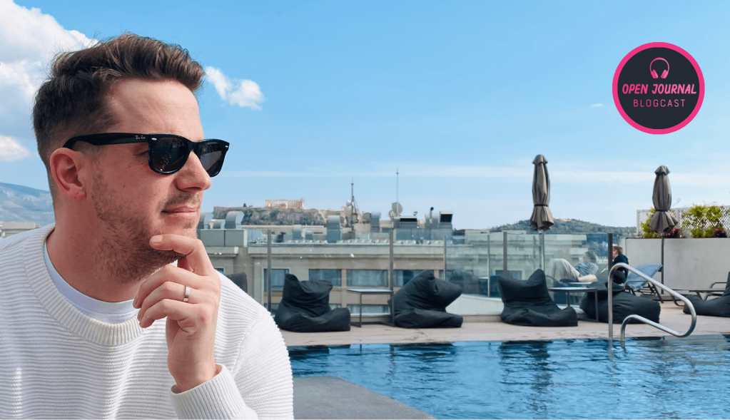 Side profile photo of Tom York looking pensively into the distance. Tom is wearing sunglasses and is resting his head on his left hand. It is a sunny day and in the distant background the Acropolis of Athens is visible.